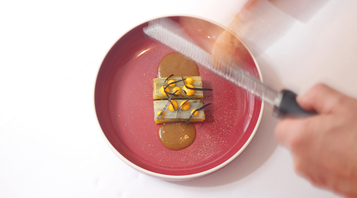 Main émiettant du pain au-dessus du plat de blettes sous forme de 2 petit pavés beiges avec sauce brune dans une assiette rose.
